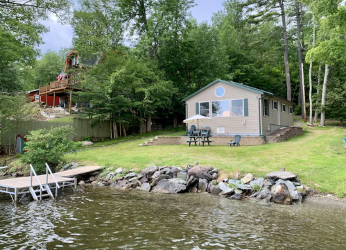 A lakeside view of a house with a patio, surrounded by trees and a small dock on the water.