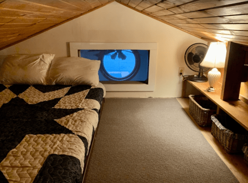 Cozy attic bedroom with a quilted bed, round window, fan, and lamp, featuring warm wooden beams and a soft carpet.