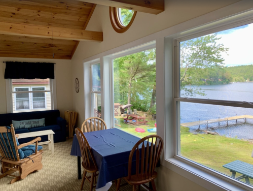 Cozy lakeside room with wooden ceiling, dining table, and large windows overlooking the water and greenery outside.