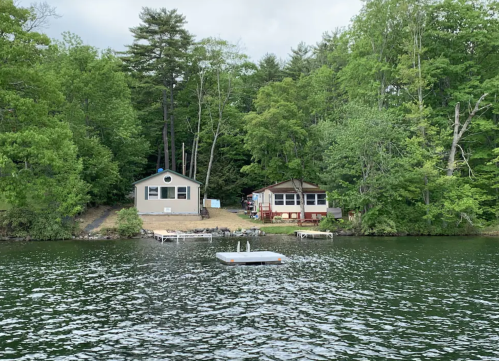 Two lakeside cabins surrounded by lush green trees, with a small dock and floating platform in the foreground.