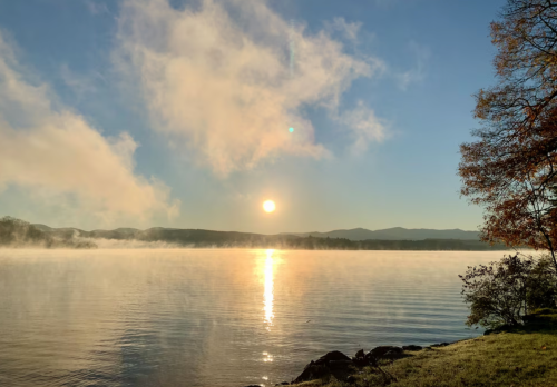 A serene lake at sunrise, with mist rising from the water and mountains in the background under a clear blue sky.