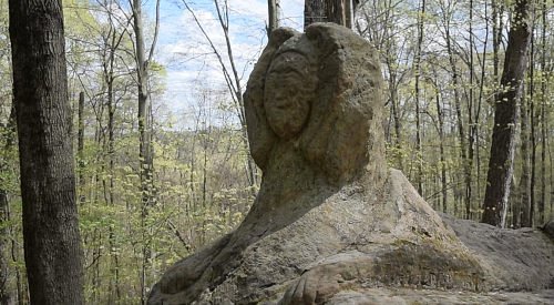 A stone sculpture resembling a figure, surrounded by trees and greenery in a natural setting.