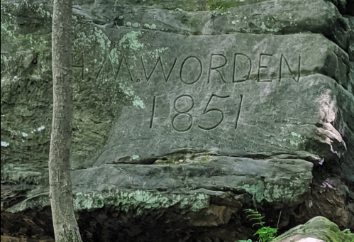 Carved inscription on a rock reading "H.A. Worden 1851," surrounded by greenery and natural rock formations.
