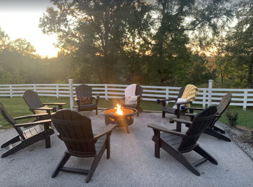 A cozy outdoor fire pit surrounded by Adirondack chairs, with a serene sunset and trees in the background.