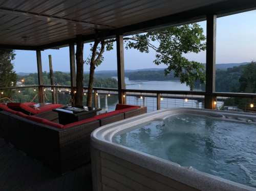 A cozy deck with a hot tub overlooking a serene river at dusk, surrounded by trees and soft lighting.