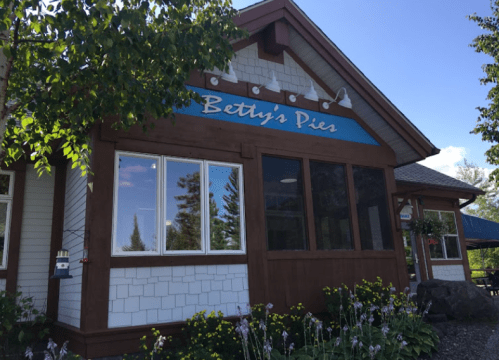 Exterior of Betty's Pies, a charming building with a blue sign, surrounded by greenery and flowers on a sunny day.