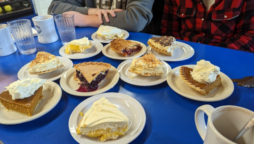 A table with various slices of pie topped with whipped cream, surrounded by glasses and coffee mugs.