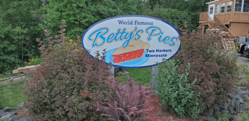 Sign for "Betty's Pies," featuring a pie graphic, located in Two Harbors, Minnesota, surrounded by greenery.