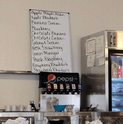 Menu board listing various pie flavors, including Apple Maple Pecan, Blueberry, and Lemon Meringue, above a drink station.
