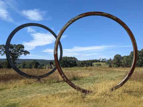 Two large metal rings stand in a grassy field, with rolling hills and a blue sky in the background.