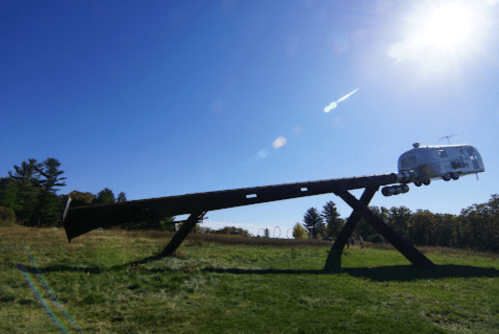 A large metal seesaw with an Airstream trailer on one end, set against a clear blue sky and bright sun.