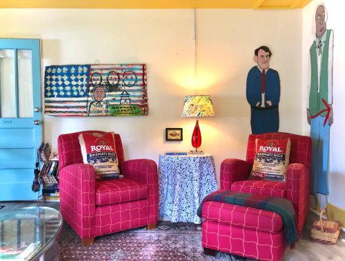 Cozy living room with red chairs, a patterned tablecloth, a lamp, and colorful wall art featuring characters and flags.