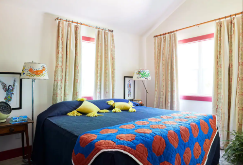 Brightly decorated bedroom with a blue bedspread, colorful pillows, and large windows with patterned curtains.