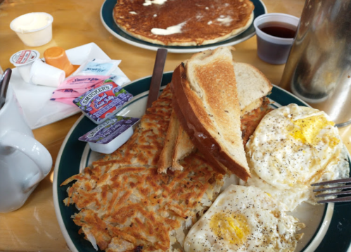 A plate of breakfast featuring eggs, hash browns, toast, and condiments, with a pancake and syrup in the background.