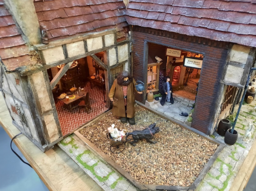 A detailed miniature scene featuring a rustic building, a figure with a cart, and a dog in a gravel courtyard.