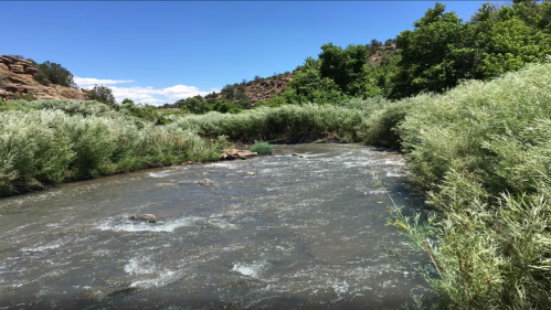 A winding river flows through lush greenery under a clear blue sky, surrounded by rocky hills.