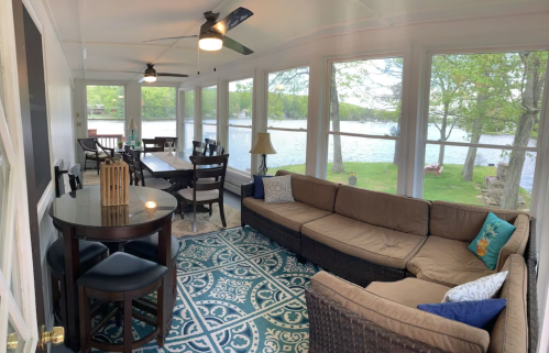 Cozy sunroom with a sectional sofa, dining area, and large windows overlooking a lake and greenery.