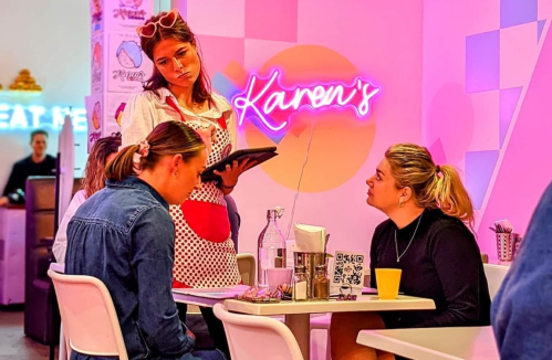 A waitress in a retro outfit takes an order from two seated customers in a colorful café with neon signs.
