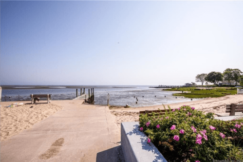 A sandy beach path leads to a calm waterway, with benches and colorful flowers in the foreground under a clear sky.