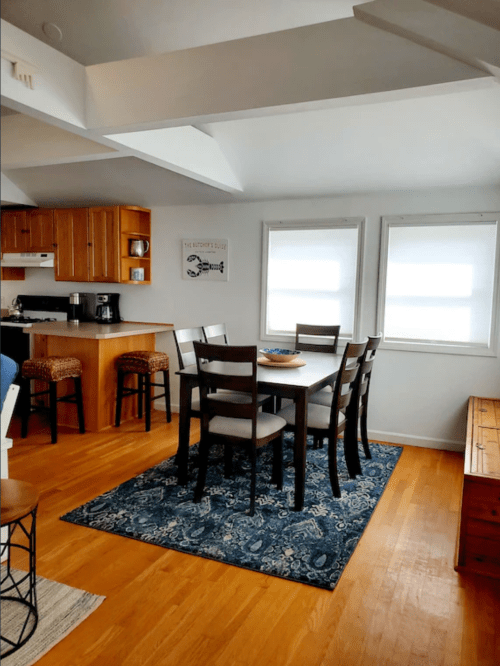 A cozy dining area with a wooden table, chairs, and a blue rug, adjacent to a kitchen with wooden cabinets.