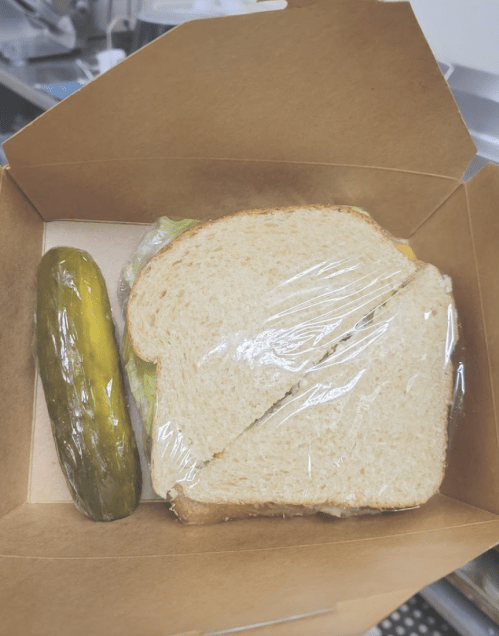A sandwich with lettuce in a takeout box, accompanied by a whole pickle on the side.