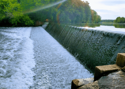 A serene river scene featuring a cascading waterfall over a dam, surrounded by lush greenery and a calm water surface.