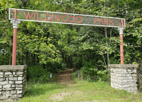 Sign for Wildwood Park at the entrance, surrounded by lush green trees and a gravel path leading into the park.