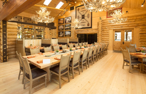 A spacious dining room with a long wooden table, antler chandeliers, and log cabin decor.