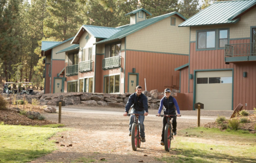 Two people ride bikes on a path near a lodge surrounded by trees and rocks.