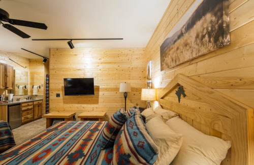 Cozy wooden bedroom with a patterned bedspread, TV, and kitchenette, featuring warm lighting and rustic decor.