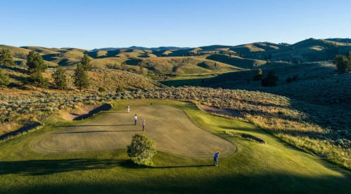 A scenic golf course with players on the green, surrounded by rolling hills and clear blue skies.