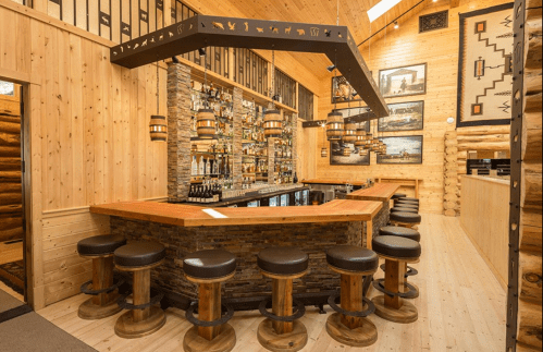 A rustic bar with wooden stools, stone accents, and a variety of bottles displayed on shelves in a cozy, warm setting.