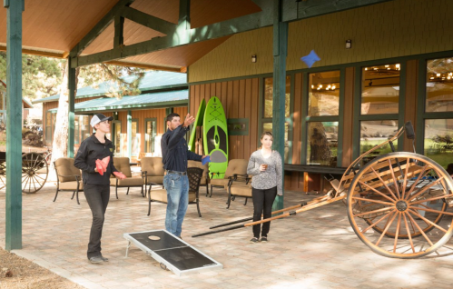 A group of three people playing cornhole outdoors under a covered patio with a wagon wheel in the background.
