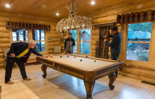 Three men play billiards in a cozy log cabin with wooden walls and a rustic chandelier.