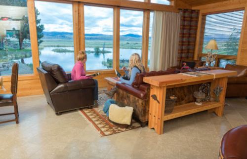 Two women sit in a cozy living room with large windows overlooking a scenic landscape.