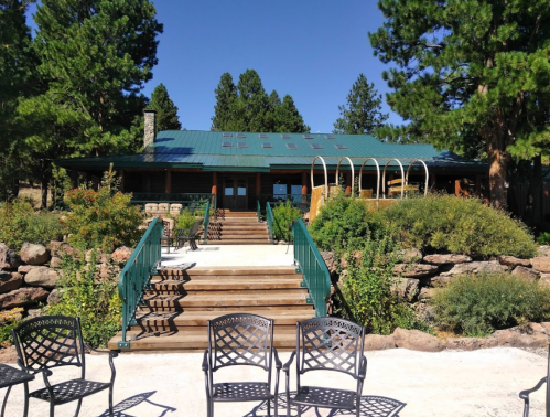 A rustic lodge with a green roof, surrounded by trees and a stone pathway leading to the entrance. Chairs are in the foreground.