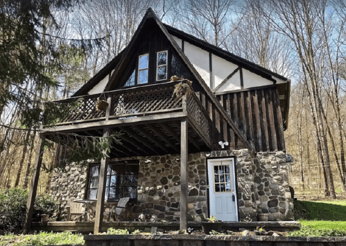 A charming two-story cabin with a stone base, wooden accents, and a balcony, surrounded by trees.