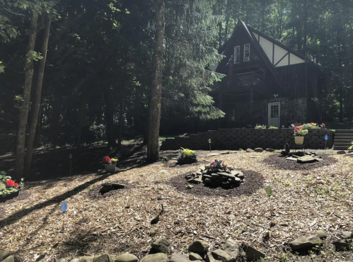 A rustic house surrounded by trees, with a landscaped garden featuring flower beds and a stone fire pit.