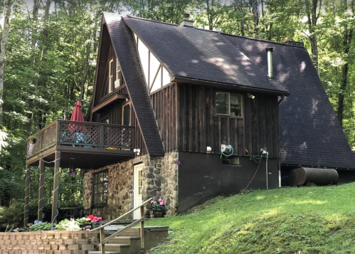 A rustic wooden house with a sloped roof, surrounded by trees, featuring a deck and stone foundation.