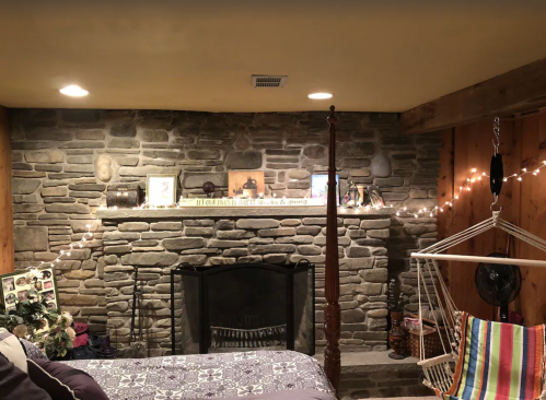 Cozy basement room with a stone wall, fireplace, and colorful hammock chair, decorated with string lights and photos.