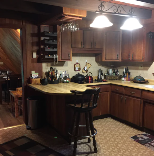 Cozy kitchen with wooden cabinets, a bar stool, and various kitchen utensils on the counter and walls.
