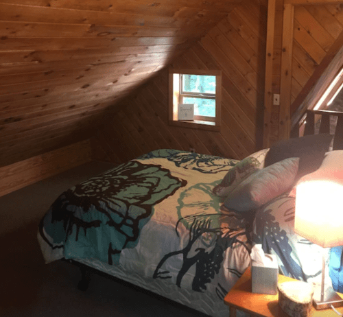 Cozy attic bedroom with a sloped wooden ceiling, floral bedding, a bedside lamp, and a small window.