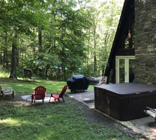 A backyard scene with a hot tub, red chairs, and a stone house surrounded by trees and outdoor decorations.