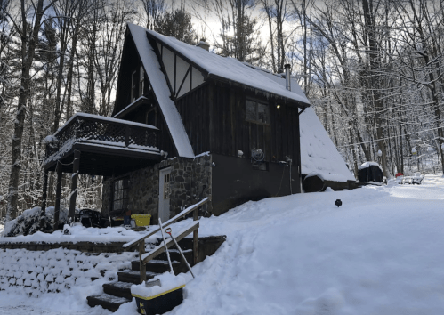 A cozy cabin in a snowy landscape, surrounded by trees, with a stone foundation and a balcony.