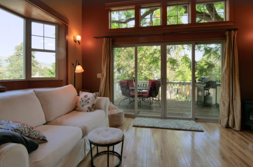 Cozy living room with a white sofa, large windows, and a view of a deck with outdoor seating and a grill.