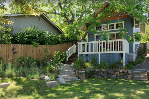 A charming blue house with a white porch, surrounded by greenery and a wooden fence, set in a sunny outdoor space.