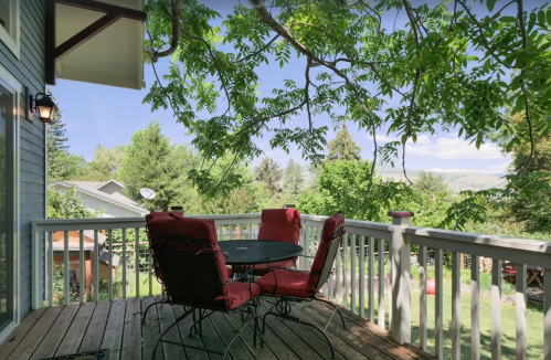 A cozy outdoor deck with a round table and red chairs, surrounded by lush greenery and a clear blue sky.