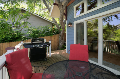 A cozy outdoor deck with a black grill, red chairs, and a view of greenery and a nearby house.