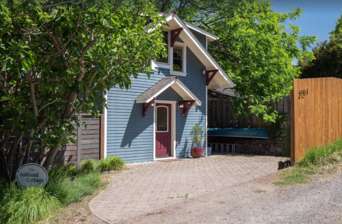 A charming blue cottage with a red door, surrounded by greenery and a wooden fence, featuring a stone pathway.