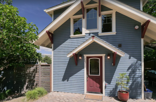 A blue two-story house with a red door and decorative roof overhangs, surrounded by greenery and a stone pathway.